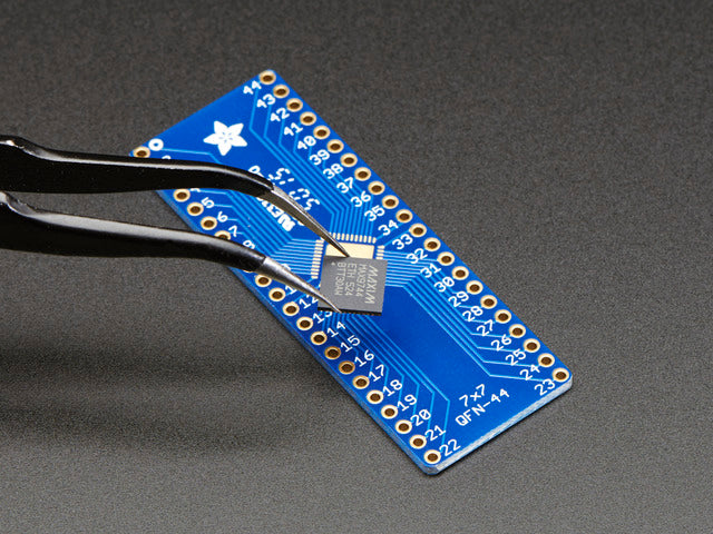 A pair of tweezers holding a microchip hovers over the SMT Breakout PCB for 44-QFN or 44-TQFP placement on a long blue rectangular breakout board.