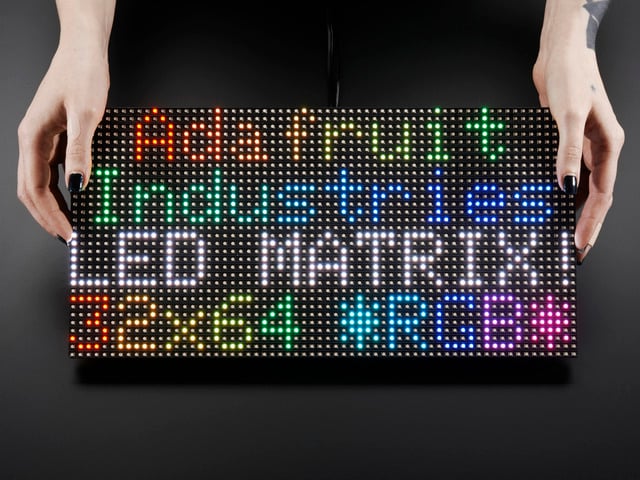 Two white hands hold out an assembled and powered on 64x32 RGB LED Matrix Panel - 6mm pitch. The matrix displays "Adafruit Industries LED MATRIX! 32x64 *RGB*"