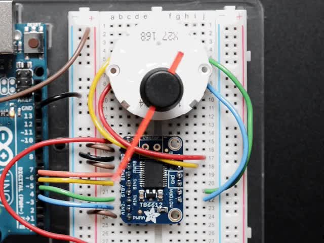 Close up of a Automotive Gauge Stepper Motor connected to a half sized white bread board, spinning. 