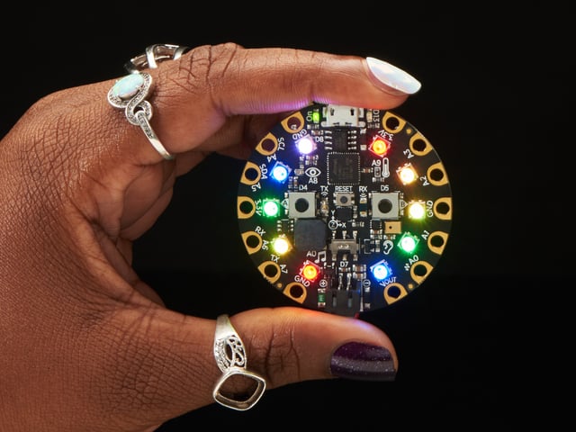 A Black woman's manicured hand holds a round microcontroller with lit up LEDs.