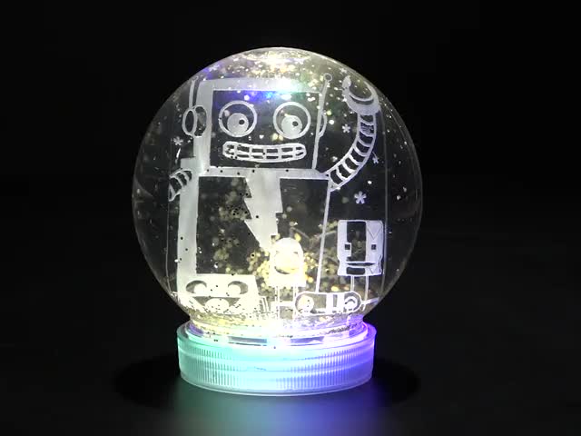 Red polished hand turning an Assembled snow globe kit with glowing electronics and friendly robot inside upside down and placing it back down. 