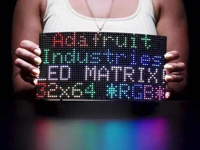 Video of woman in a white sleeveless t-shirt holding and slightly bending a black flexible 64x32 RGB LED Matrix. The LED matrix is lit up with rainbow LEDs with the text: Adafruit Industries LED Matrix 32x64 *RGB*.