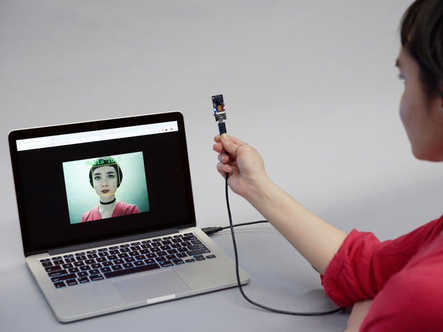 Angled shot of a ESP-EYE Development Board connected to a Macbook and being held by a female facing the camera toward themselves. 