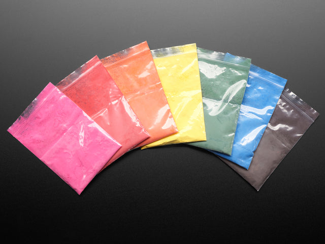 Angled shot of six clear baggies fanned out. The bags contain powder paint pigments separated by color: red, orange, yellow, green, blue, black, and pink.