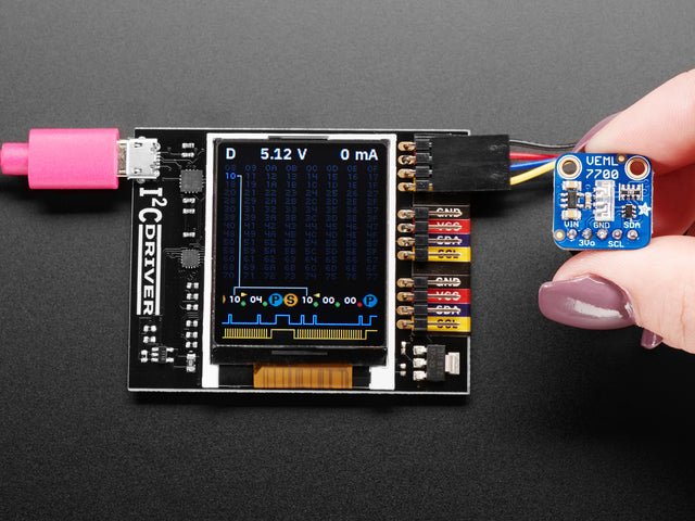 Top down view of a  I2CDriver by Excamera Labs attached to a small blue squared breakout board being held by a white hand. 