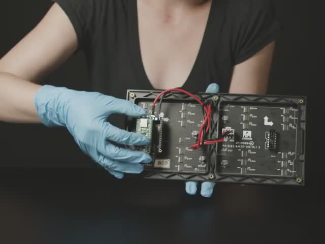 White woman wearing blue gloved rotating an RGB LED matrix. The LED animation resembles falling colored sand.