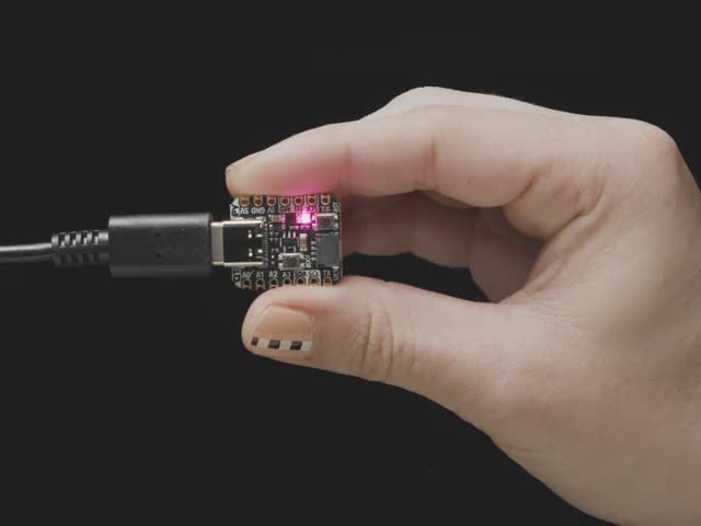 Video of hand holding a QT Py PCB in their hand. An LED glows rainbow colors.