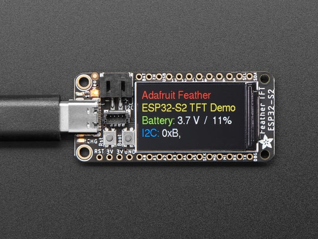 Adafruit ESP32-S2 TFT Feather powered on by a USB- C power source displaying the product tittle in a red, yellow, green, white and blue. 
