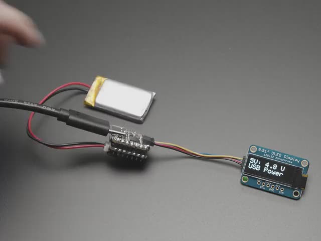 Video of a person with white painted nails unplugging a USB cable from a small, black, square-shaped lipo battery breakout board soldered to a similarly shaped microcontroller, which is also connected to a monochrome OLED display breakout. The OLED breakout displays battery and power data.