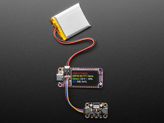 Top view of a purple rectangular microcontroller with a color TFT screen and powered by an ESP32-S3 chip. It is also connected to a Li-Poly battery and a lux sensor breakout. The TFT display reads in colored text: "Adafruit Feather

ESP32-S3 TFT Demo

Battery: 3.8V / 45%

I2C: 0xB, 0x10, "