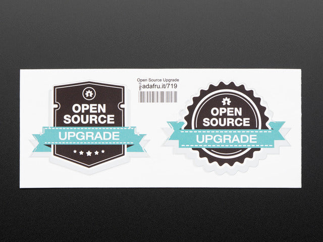photograph of two black stickers, one shield shaped, the other gear shaped, with white lettering reading "open source" with a turquoise banner running across each reading "upgrade" both stickers are mounted on white paper with barcode. 